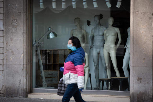 Una mujer protegida con una mascarilla pasa junto a un escaparate de una tienda cerrada a consecuencia del coronavirus, en Barcelona/Catalunya (España) a 26 de marzo de 2020.