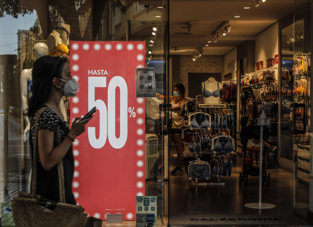 Una mujer protegida con mascarilla pasa al lado del escaparate de una tienda donde se observan carteles indicativos de rebajas en Valencia.