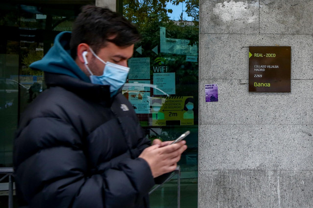 Un joven mira su móvil delante de una sucursal bancaria en Collado Villalba, Madrid (España)