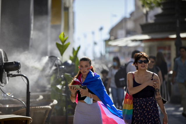 Archivo - Dos chicas con un bolso y una bandera LGTBI, al lado de un difusor de vapor de agua de un restaurante de la calle Alcalá, a 2 de julio de 2021, en Madrid, (España).