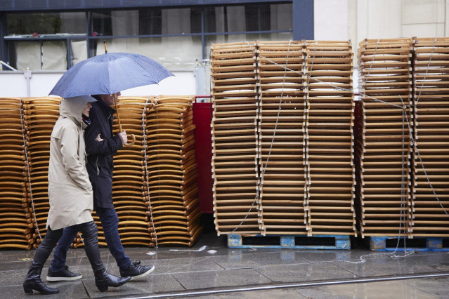 Archivo - Una pareja pasea bajo su paragüa con las sillas apiladas de fondo durante la vuelta de la lluvia a Sevilla en Semana Santa