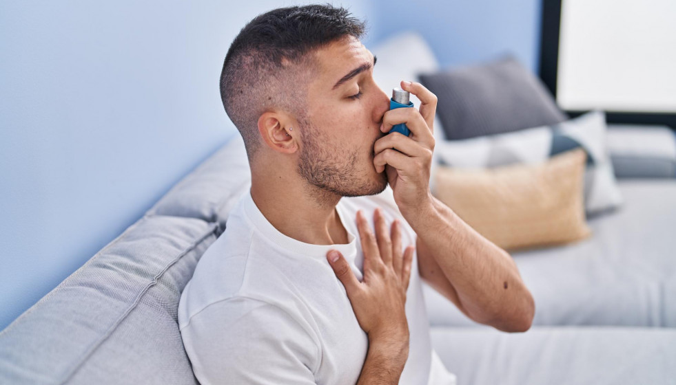 Young hispanic man using inhaler sitting sofa home
