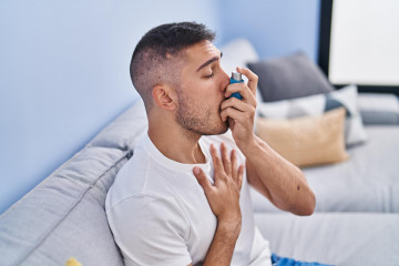 Young hispanic man using inhaler sitting sofa home