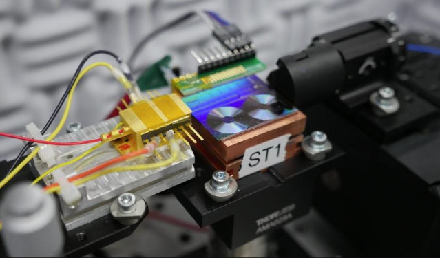 Archivo - Investigadores del NIST prueban un chip para convertir la luz en señales de microondas. En la foto aparece el chip, que es el panel fluorescente que parece dos pequeños discos de vinilo. La caja dorada  es el láser semiconductor que emite luz al