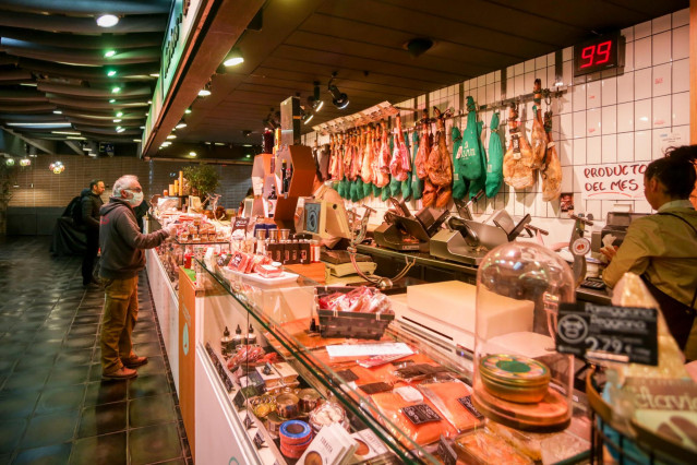Archivo - Varios clientes hacen la compra en un puesto de carne y embutidos en el mercado de San Antón, que permanece abierto y anuncia que también atiende a domicilio, en Madrid (España), a 18 de marzo de 2020.