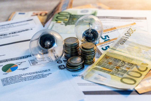 Archivo - Dos bombillas sobre billetes, monedas y facturas en imagen de archivo.